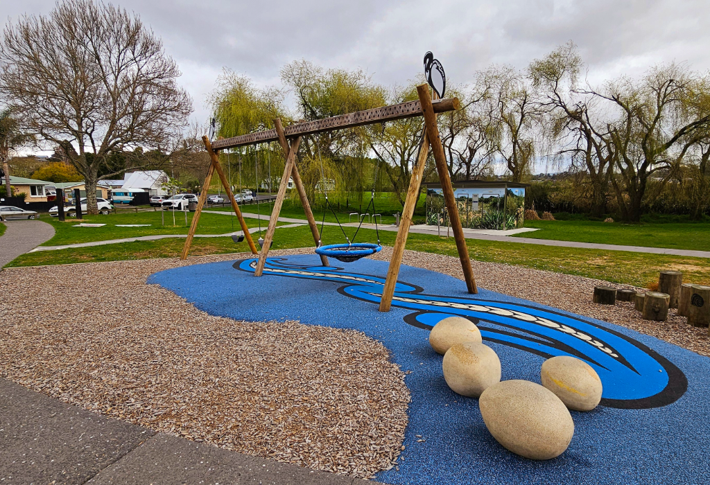 Level concrete path leads to a timber swing set with a basket swing. Surfaces are blue rubber softfall with an eel motif and wood chips. Features four large egg-shaped stone seats.