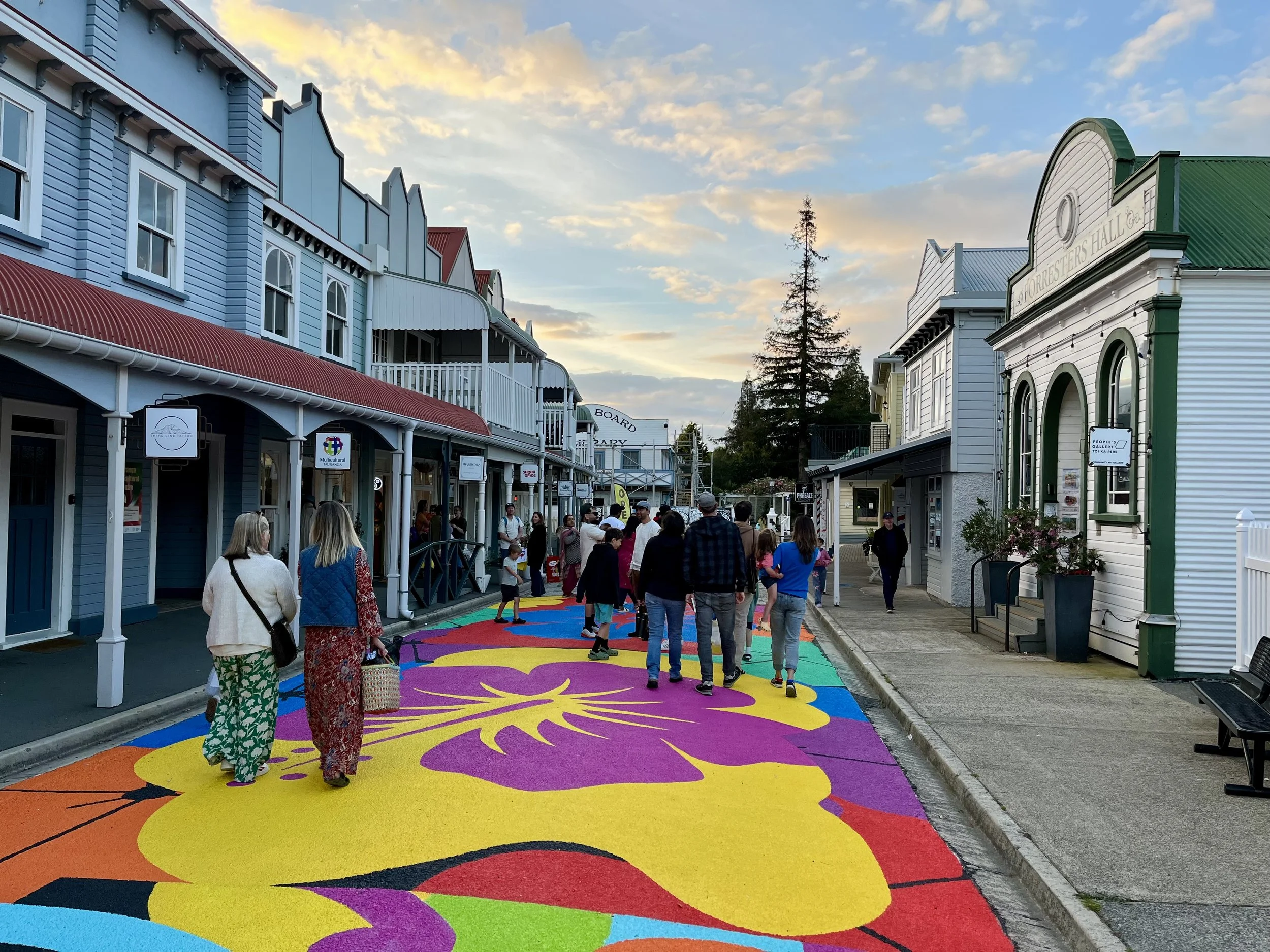 Wide, level concrete walkway featuring a vibrant multi-coloured floral mural. A black metal bench sits on the right. Level shop entrances are located under covered timber awnings.