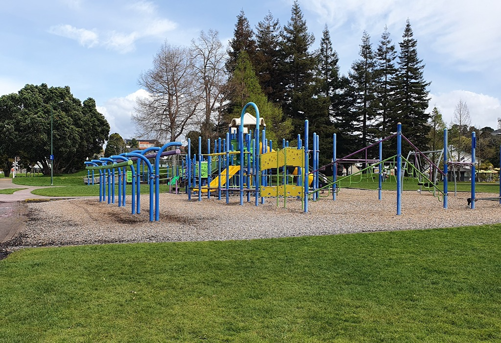 Blue and yellow playground equipment on a level woodchip surface. A flat paved pathway leads to the area, which is surrounded by an expansive grass lawn and tall evergreen trees.