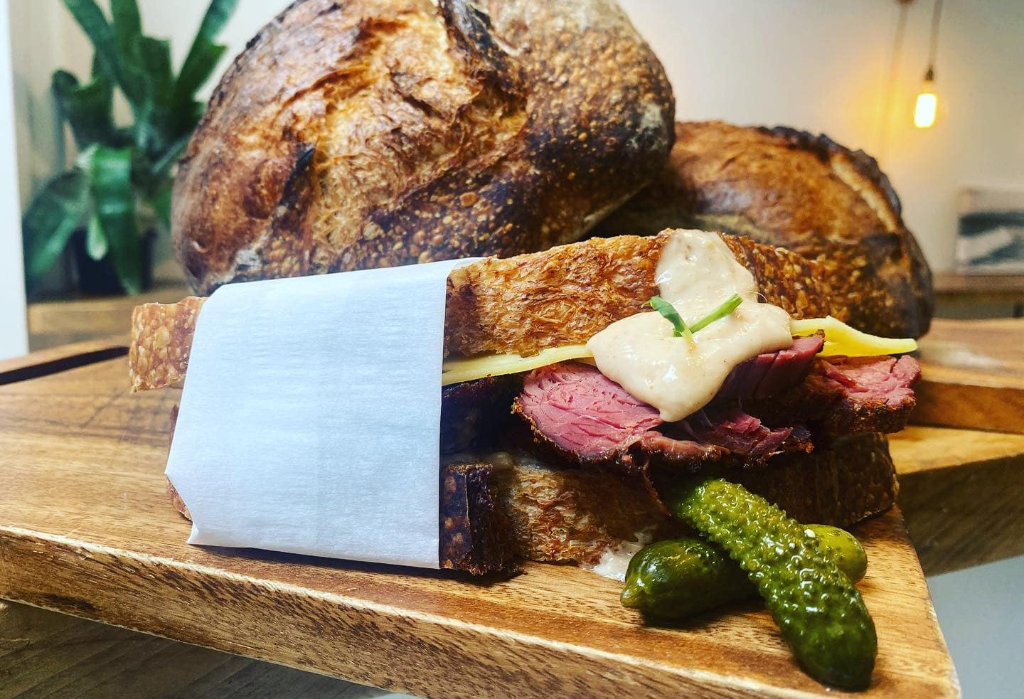 Pastrami sandwich on thick sourdough bread with cheese and creamy sauce on a flat wooden board. Two gherkins are on the side, with large dark-crusted loaves in the background.