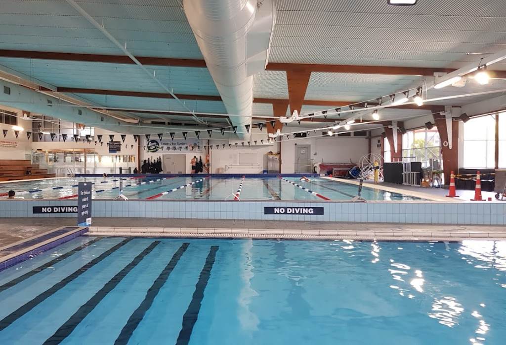 Indoor pool with blue tiled steps and dark contrast strips. A lap pool sits behind a level grey tiled edge with 'NO DIVING' signage. Large windows and track lighting provide bright illumination.