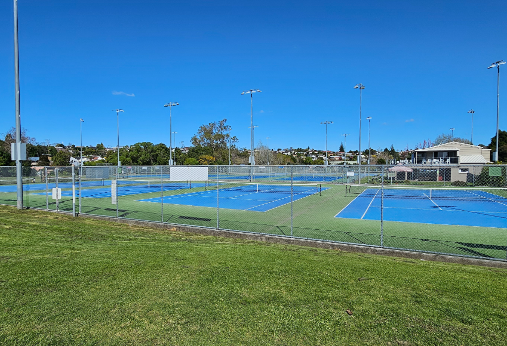 Blue hard-court tennis courts with white markings and a level gate in a wire fence. A concrete kerb borders the courts next to sloped grass. Tall light poles and a clubhouse in background.