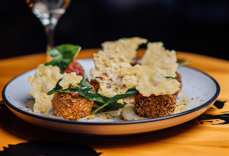 Golden-brown fried balls topped with green leaves and thin yellow crisps on a white plate with a dark rim, set on a yellow table surface.