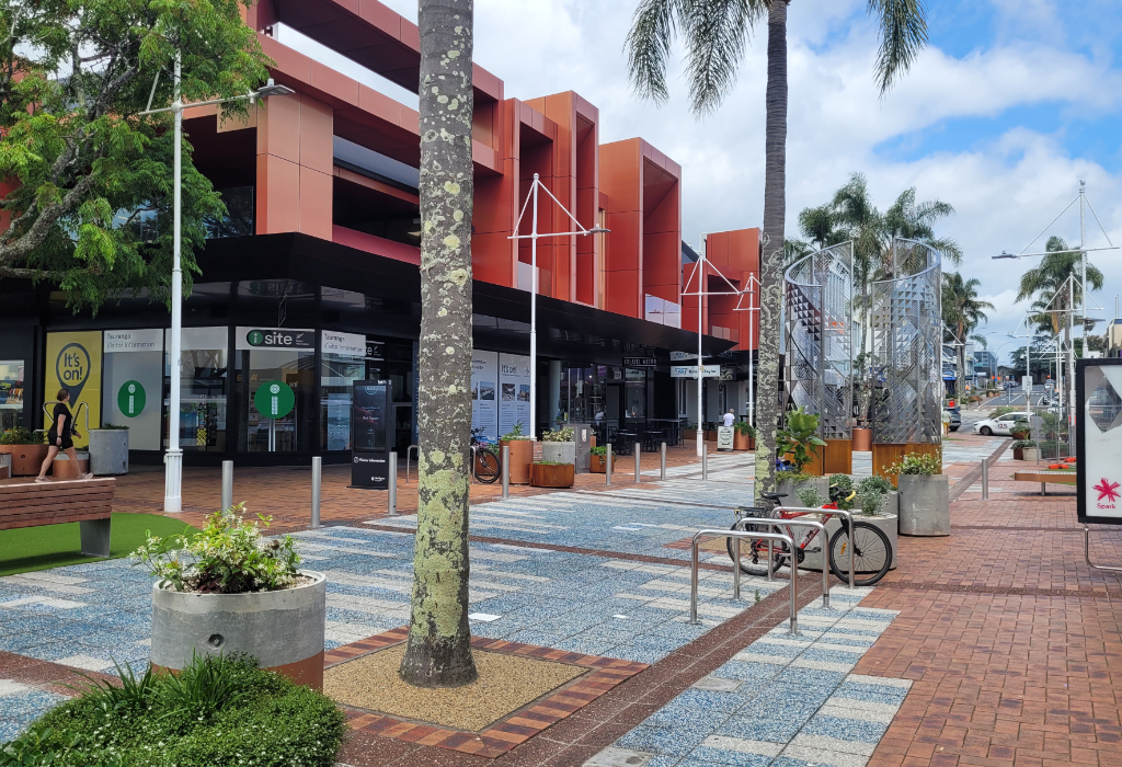 Level paved plaza with blue and grey tiles leads to a flat entrance for an 'i-site' visitor centre. Includes wooden benches, bike racks, and wide pathways around concrete planters and palm trees.