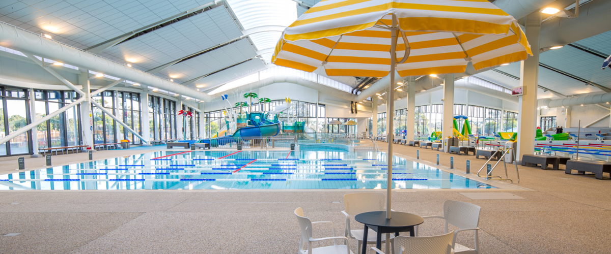 Wide, level, speckled non-slip deck with metal handrails and ramped pool access. Bright lighting over lap pools, water play area, and cafe seating under a yellow and white striped umbrella.