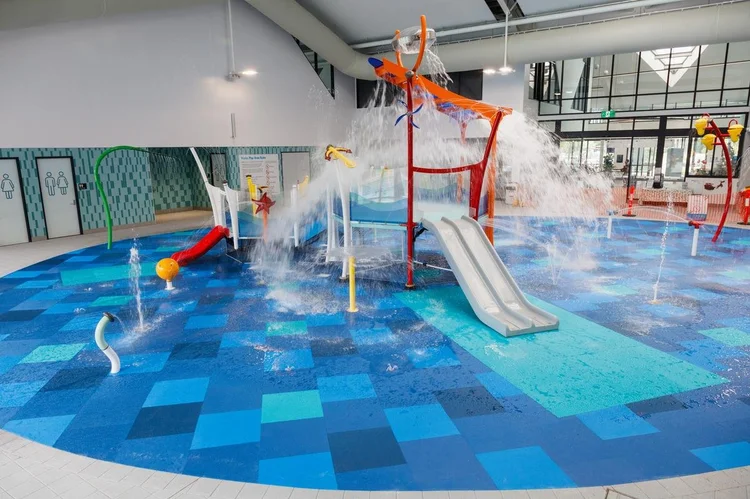 Level indoor splash pad with blue patterned tiles. Orange play structure with grey slide and water jets. Restroom doors with symbols on the left. High ceiling with bright overhead lighting.