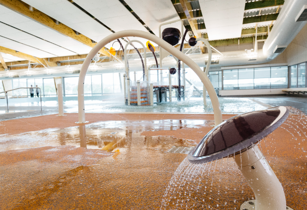 Level indoor splash pad with textured orange non-slip surfacing. Features white spray arches, a mushroom fountain, and shallow pool access with metal handrails. Brightly lit by large windows.
