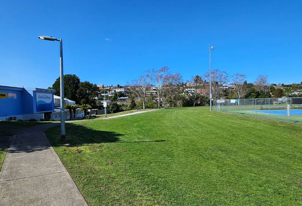 Level concrete path leads past a blue building with an 'Ōtūmoetai Pool' sign. A wide grass field is adjacent to fenced blue tennis courts. Tall grey light poles stand near the pathways.