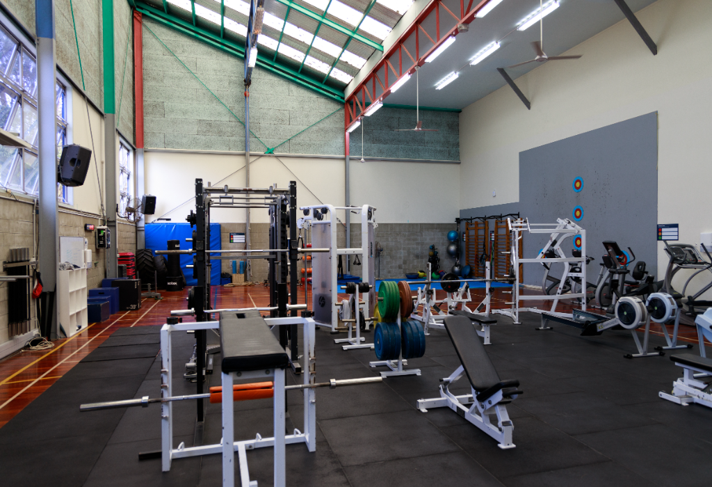 Spacious gym with level dark rubber mats on a timber floor. Features weight racks, benches, and cardio equipment. Brightly lit by skylights and overhead lights with wide, level pathways.