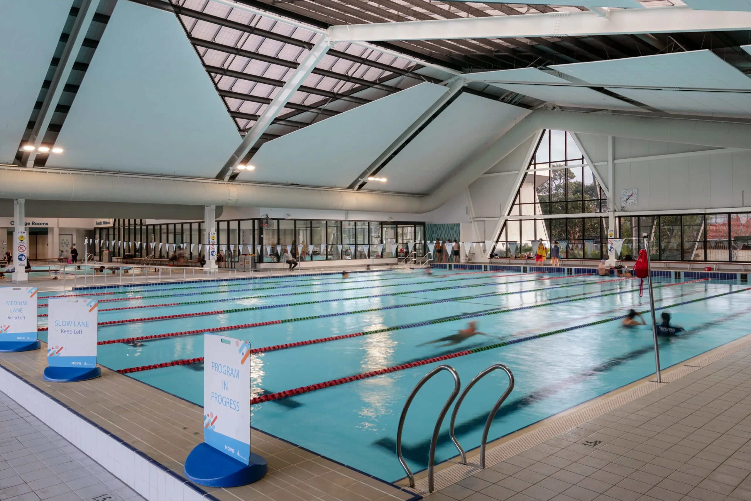Level light grey tiled floor surrounds an indoor pool. Signs denote "Slow Lane", "Medium Lane", and "Program in Progress". Curved stainless steel handrails lead into water. High glass ceiling.