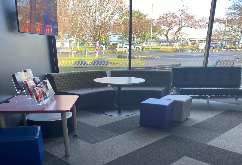 Level grey carpeted seating area with low sofas, cube ottomans, and a round table. Floor-to-ceiling windows provide natural light and views of a level outdoor paved area.
