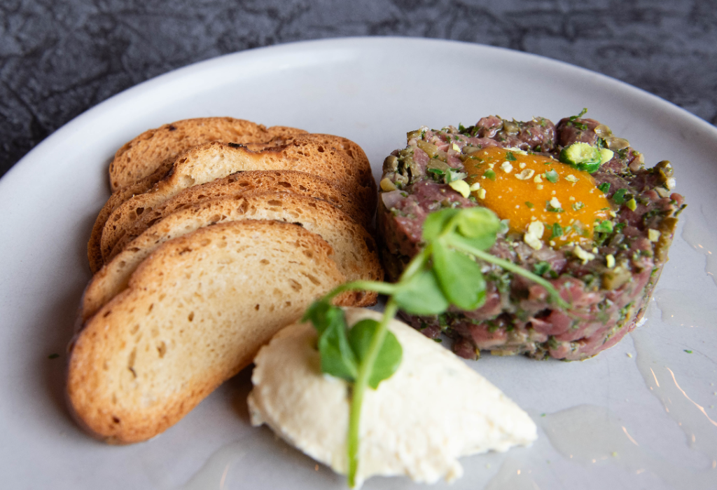 Circular mound of beef tartare topped with a yellow yolk, served on a white plate with four toasted bread slices, a scoop of white cream, and a green pea shoot garnish.