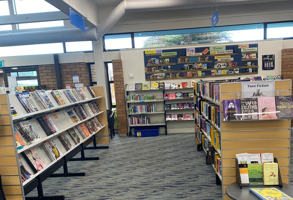 Level library area with wide pathways on blue-grey patterned carpet. Shelving units for books and magazines are clearly labelled with high-contrast signage under bright overhead lights.