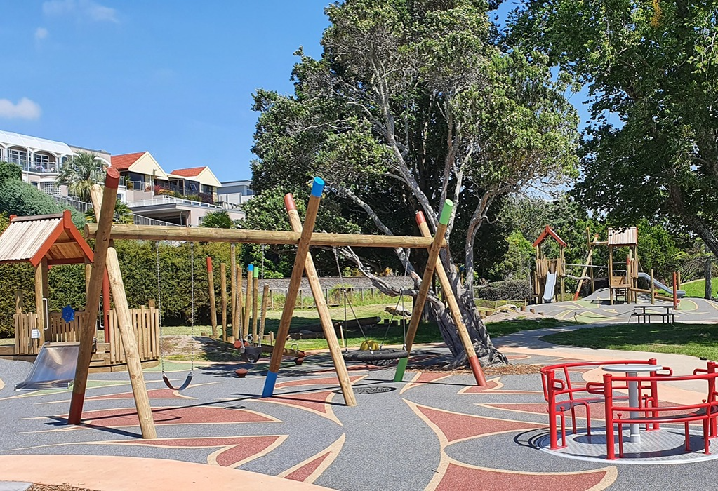 Level playground with multicoloured patterned rubber safety matting. Includes a wooden swing frame, red carousel, and wide concrete pathway winding between large trees and seating.