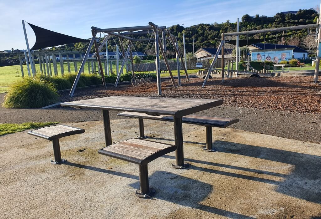 Wooden-topped picnic table on a level concrete surface with black metal legs. One long bench and two square seats are provided, with an open end suitable for wheelchair access.