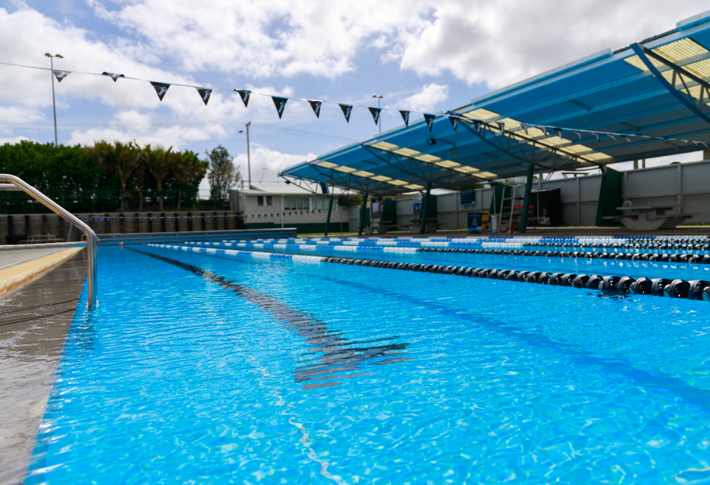 Outdoor pool with lane ropes. A silver metal handrail is at the water's edge next to a level grey concrete deck. A blue sheltered area with benches and overhead flags is in the background.