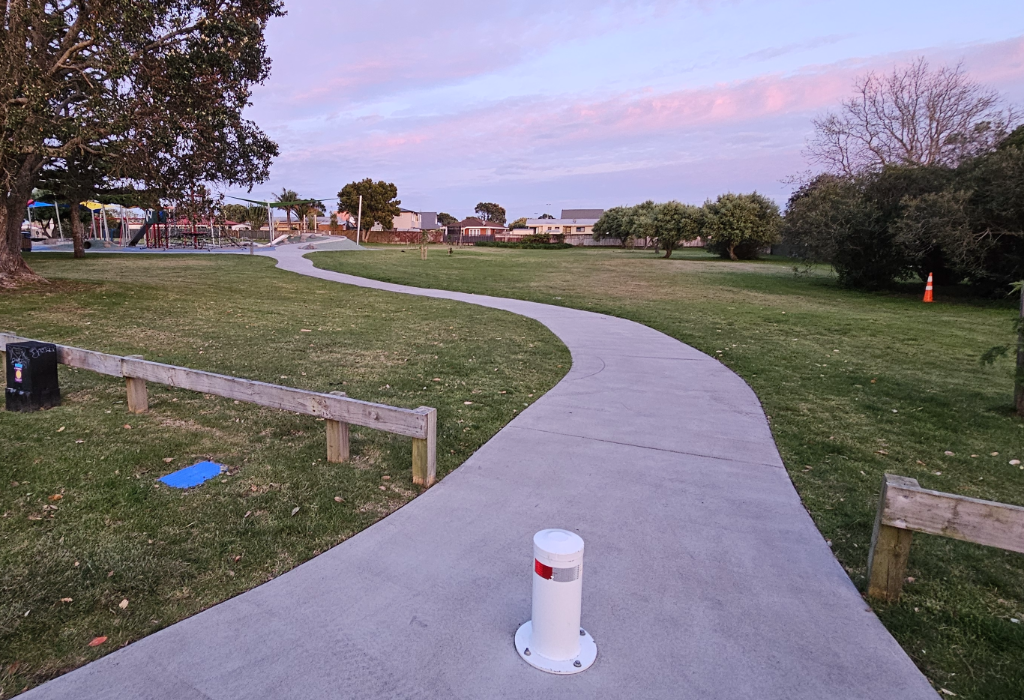 Wide, level concrete path winds through grass. A white bollard sits in the centre of the path entrance between low timber fences. Playground and shade sails in the distance.