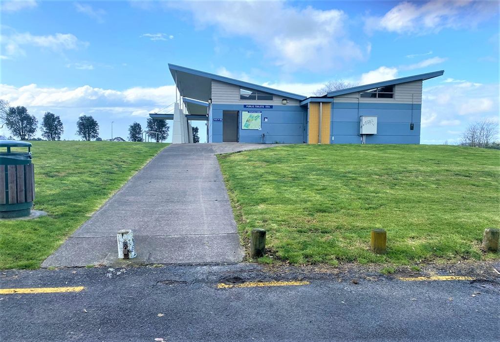 Wide concrete pathway with a gentle gradient leads across grass to a light blue public toilet block. A map and signage are visible on the exterior wall near the doorway.
