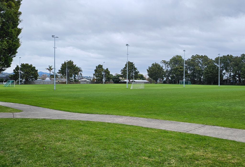 Wide level concrete pathway across a flat green lawn leading to a large grass sports field with white goals and tall floodlight poles. All surfaces appear smooth and unobstructed.