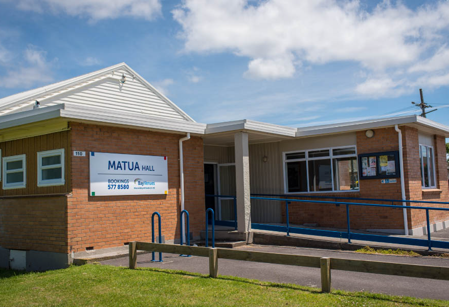 Matua Hall entrance via concrete ramp with blue handrails. Asphalt path and timber fence in foreground. Red-brown brick walls with large signage and a small wheelchair symbol next to windows.