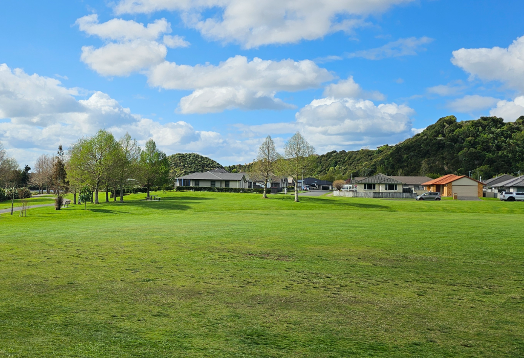 Level green lawn with a flat, grey paved pathway. A timber park bench provides seating near a row of trees. Houses and green hills border the space under a bright blue, cloudy sky.
