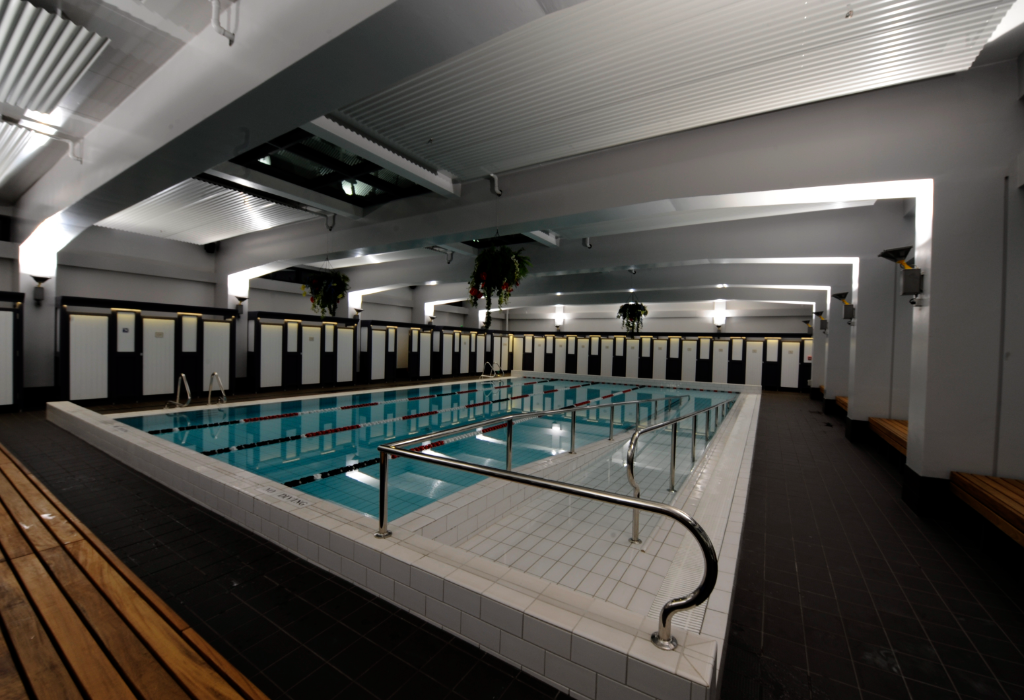 Indoor pool with a white tiled access ramp and silver handrails. Dark tiled level floor with wooden benches. White changing stalls line the back wall under bright overhead lighting.