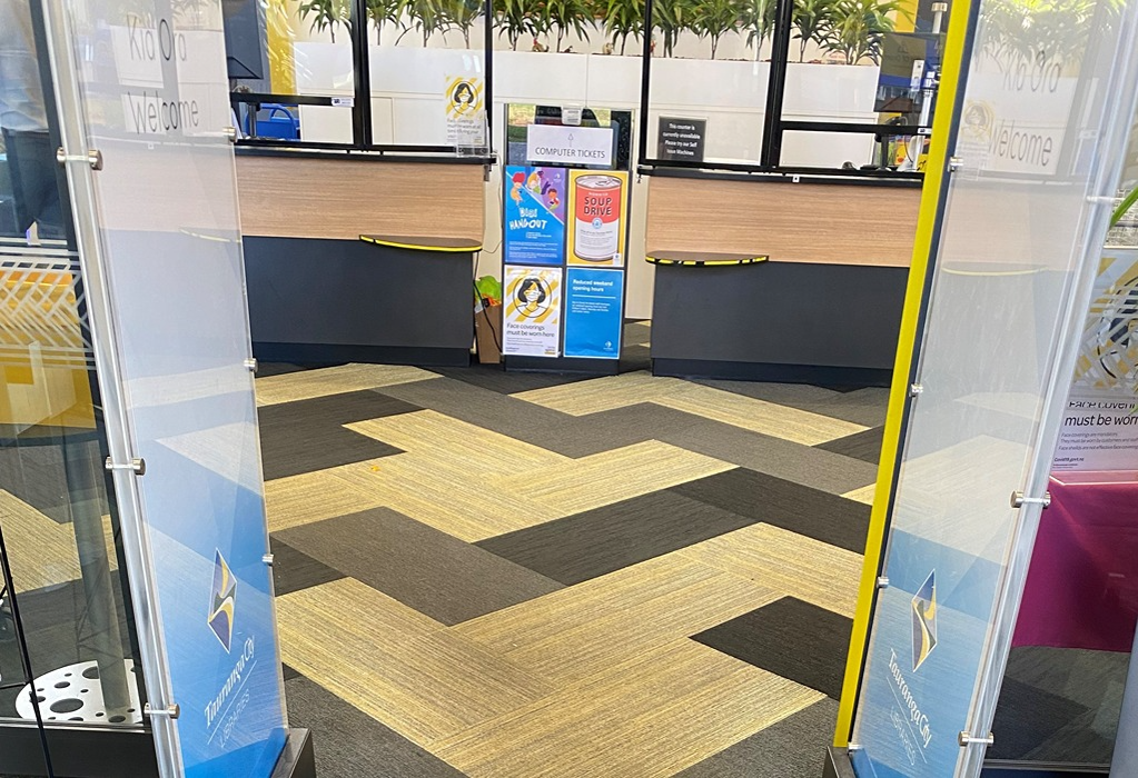 Wide level entrance to Tauranga City Libraries through glass panels. Flat patterned carpet in grey and beige. Two service counters with clear screens and a lower yellow shelf in background.