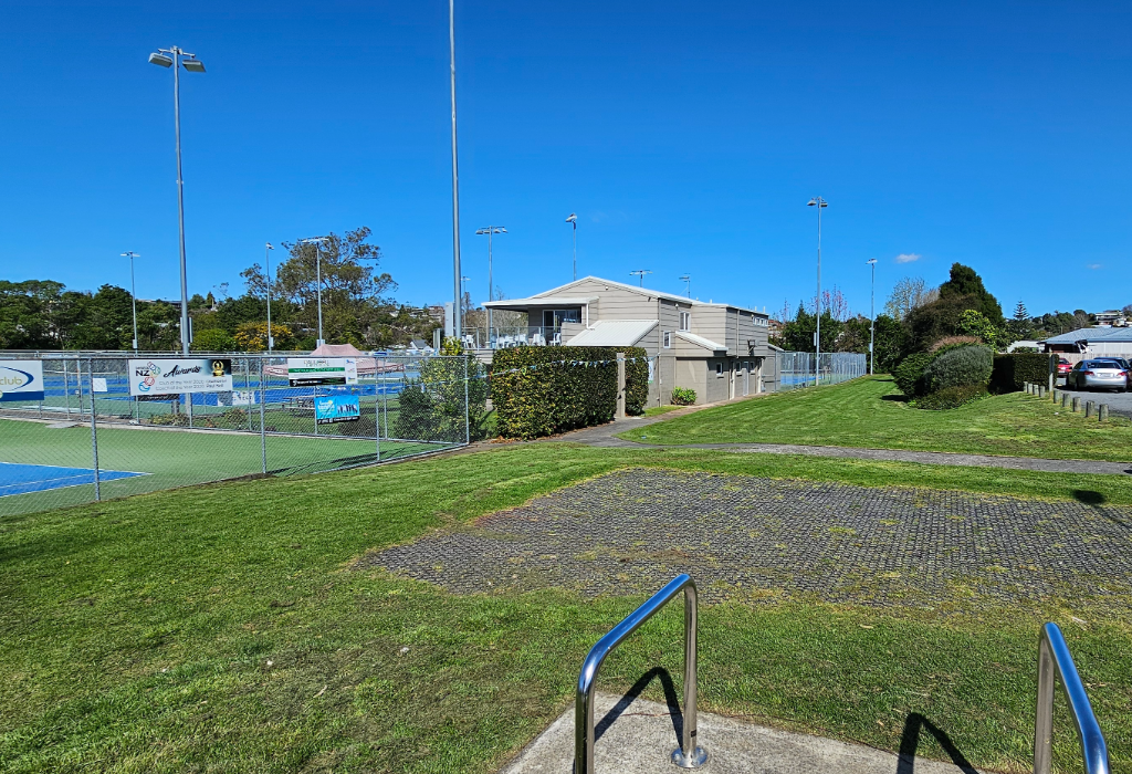 Metal handrails on concrete overlook a mesh-reinforced grass area. A level concrete path leads to a beige building with a ramped deck, situated beside fenced blue tennis courts.