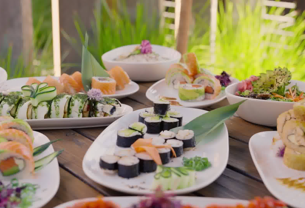 Dark timber outdoor table with level, horizontal planks, holding multiple plates of sushi and salad bowls. Blurry green foliage and a timber fence in the background.