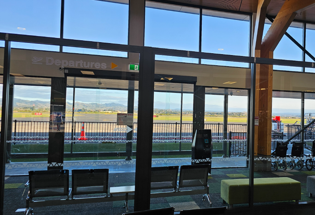 Level carpeted waiting area with metal seating. A large glass automatic door marked 'Departures' leads to a level outdoor walkway. Natural light enters through floor-to-ceiling windows.
