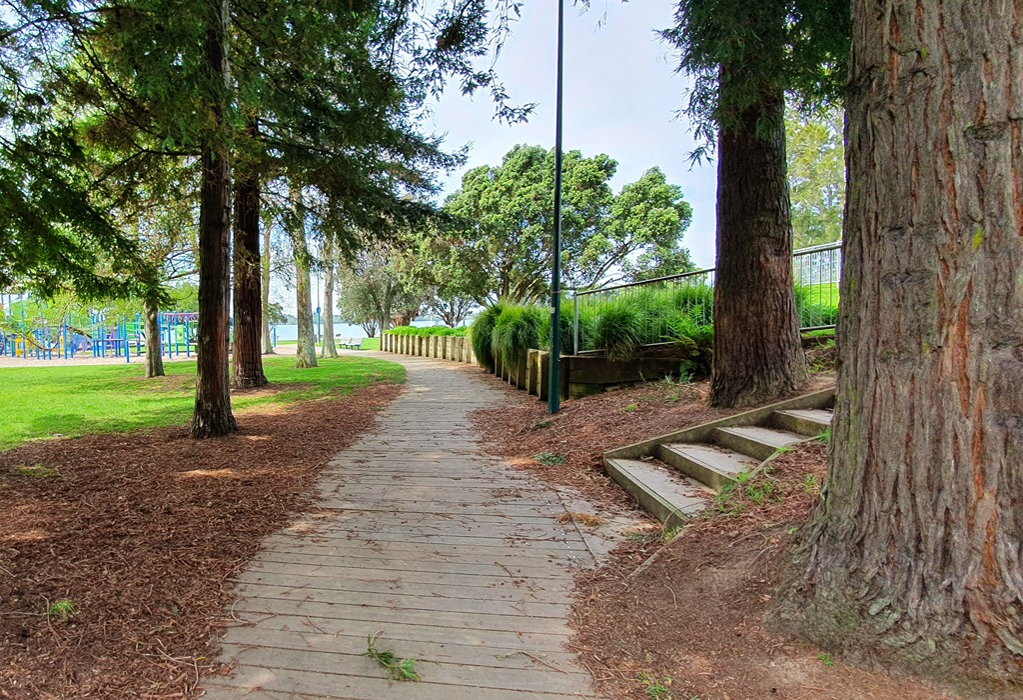 Wide timber boardwalk curves past large trees. Short concrete stairs lead uphill on the right. A metal handrail borders a raised garden bed with a timber sleeper retaining wall.