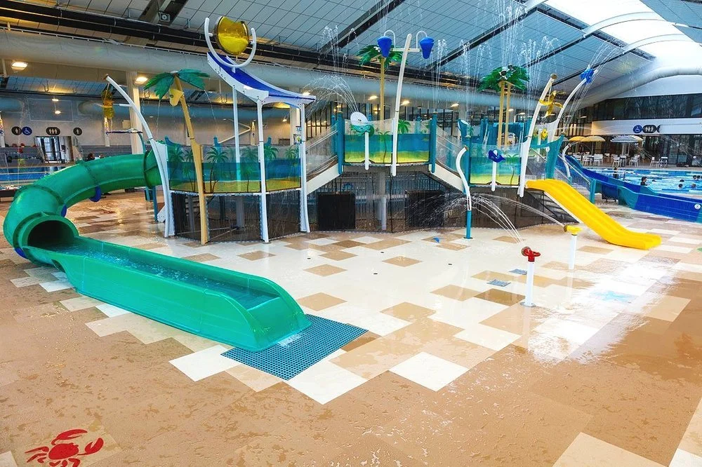 Level tan and cream tiled floors. Multi-level play structure with ramped access, a green tube slide, and a yellow slide. Blue accessibility signage and level pool entry visible in the background.