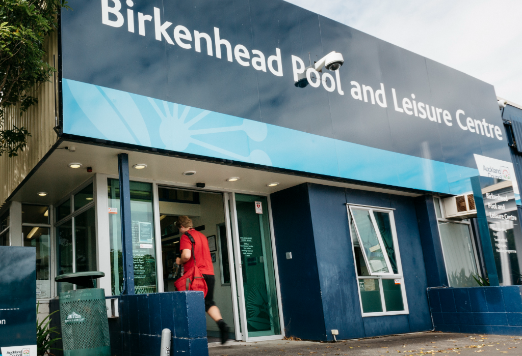 Level concrete entrance to Birkenhead Pool and Leisure Centre with wide glass doors, overhead recessed lighting, and large blue and white signage.