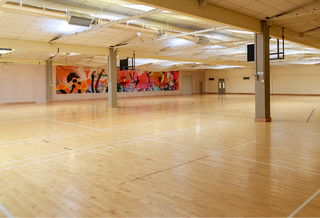Wide, level indoor court with polished light timber flooring and white markings. Thick support columns, bright overhead lighting, and a double door entrance visible at the far end.