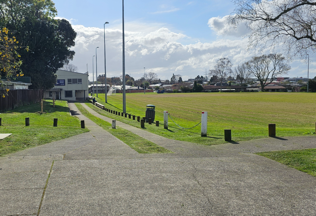Level concrete pathways fork towards a white two-storey building and a large grass field. Low wooden bollards and tall light poles line the wide, flat paths under a cloudy sky.