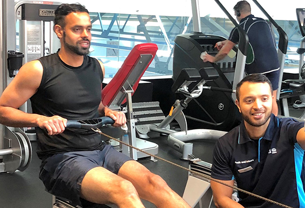 Well-lit gym with level, dark grey flooring. Wide, clear floor space between a low rowing machine, red weight bench, and other exercise equipment.