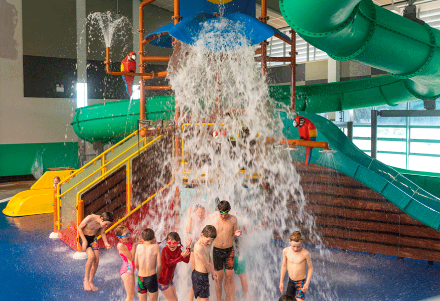Indoor water play area with blue flooring. Yellow stairs with handrails lead to a platform and green slide. Water spills from a central bucket feature onto children. Bright indoor lighting.