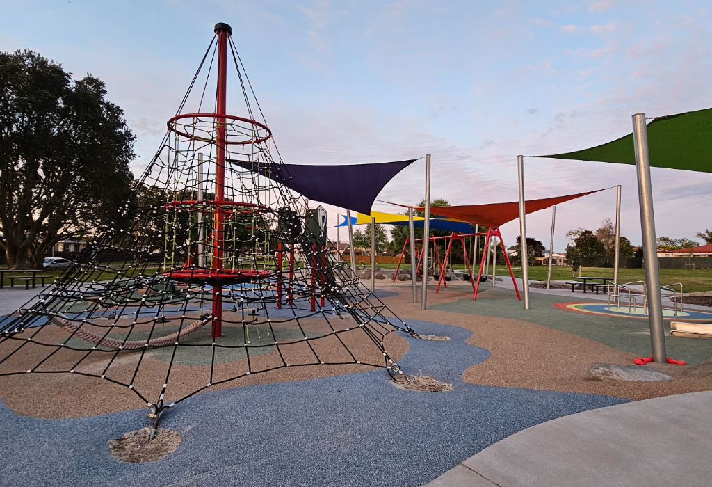 Level playground with blue and tan rubber soft-fall surfacing and smooth concrete paths. Features a large rope climbing net, red swings, and colourful shade sails supported by silver metal poles.