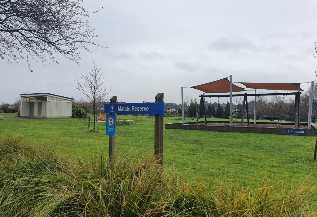 Blue Matatu Reserve sign in a flat grassy park. Cream utility building with level entry. Swing set under brown shade sails with a low timber border. No paved pathways visible on the grass.