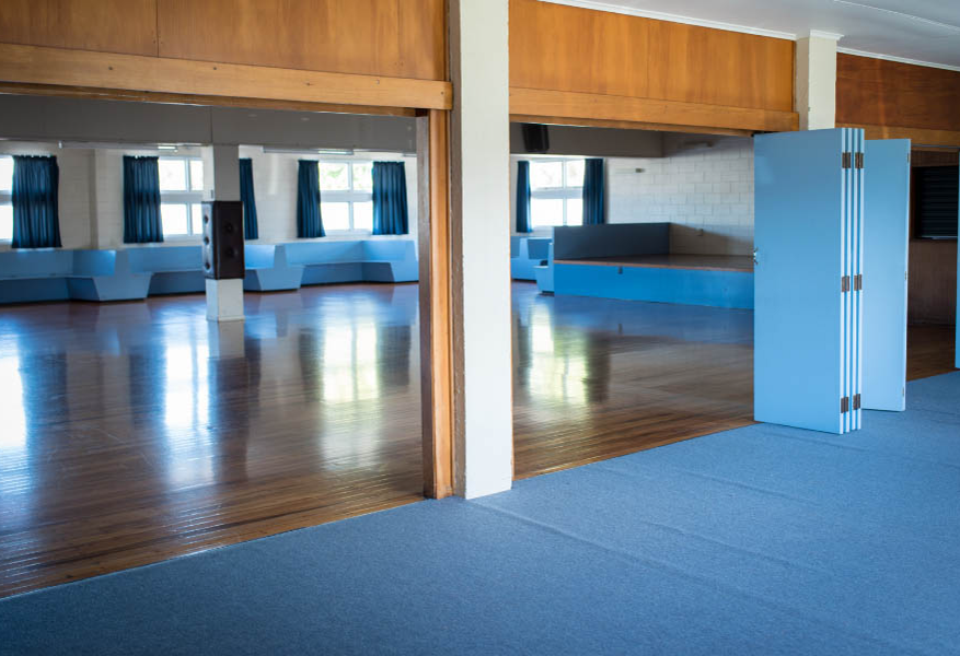 Wide, level entrances separated by a pillar lead from blue carpet to a polished timber floor. Light blue bench seating and a raised platform line the walls below blue-curtained windows.
