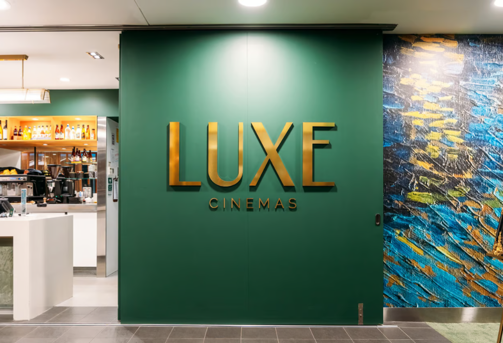 Large gold 'LUXE CINEMAS' signage on a green wall. Level dark grey tiled flooring leads through a wide, brightly lit opening on the left to a cafe area with white counters.