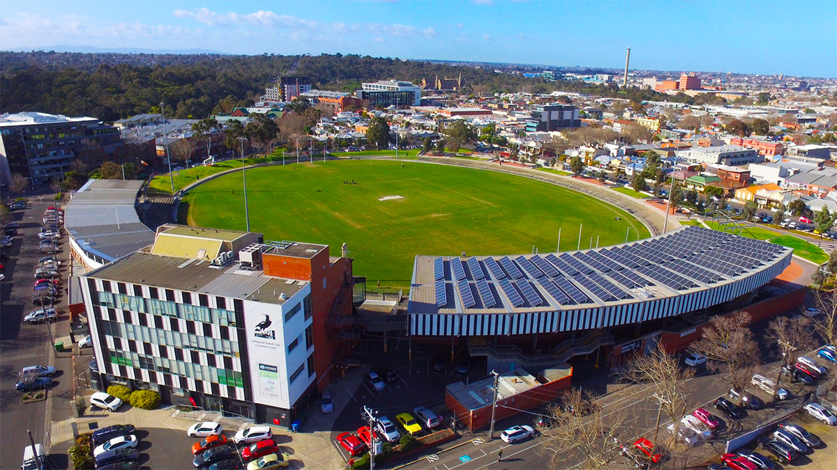 Level street access to multi-storey centre and curved grandstand. Asphalt car parks, concrete footpaths and stadium lighting surround a green grass oval and brick buildings with solar panels.