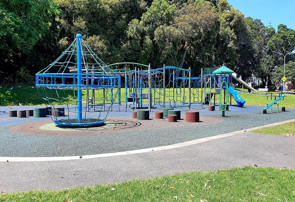 Level asphalt pathway curves around a playground with blue and grey rubber soft-fall surfacing. Includes blue metal climbing structures, a slide, and various low cylinder-shaped seating blocks.