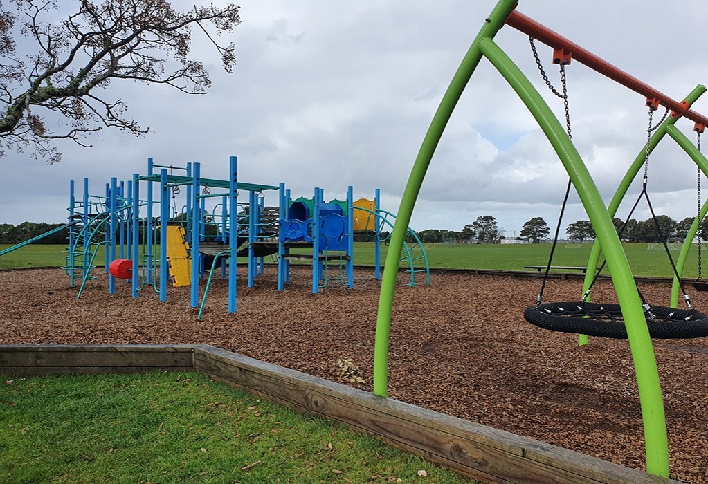 Blue and yellow climbing frame and lime green swing set with a black net seat on wood chip bark. Low timber sleeper border separates equipment from grass. Open field and trees in the background.