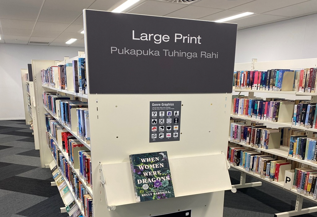 "Large Print" sign in white on dark grey. White metal shelving on level grey and black patterned carpet. Bright overhead lights and wide, level walkways between rows of book stacks.