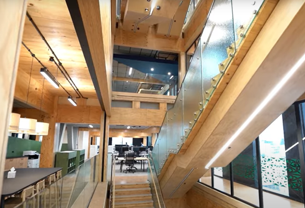 Level timber floor leads to a wide wooden staircase with glass balustrades and metal handrails. Bright LED lighting under stairs. Open-plan office area with desks and chairs in background.