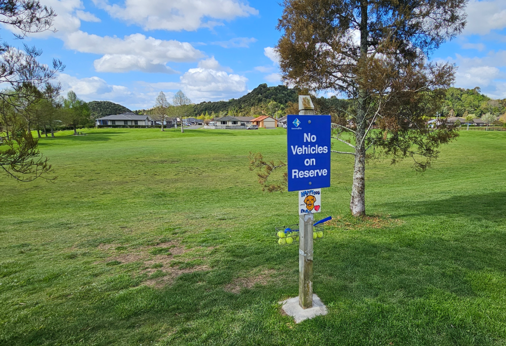 Blue 'No Vehicles on Reserve' sign on a wooden post. Wire basket with yellow tennis balls attached below. The terrain is a level, short green grass surface with no paved pathways.