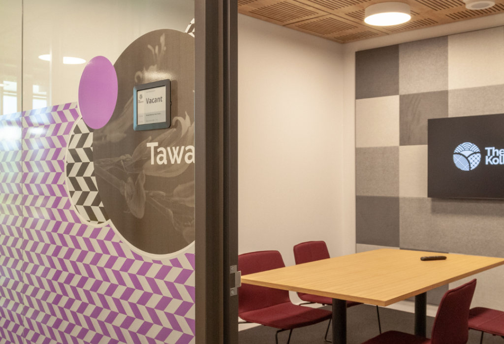 Level entrance to a meeting room with glass partition and digital 'Vacant' sign. Inside features a light timber table, burgundy chairs, and a carpeted floor under warm ceiling lights.