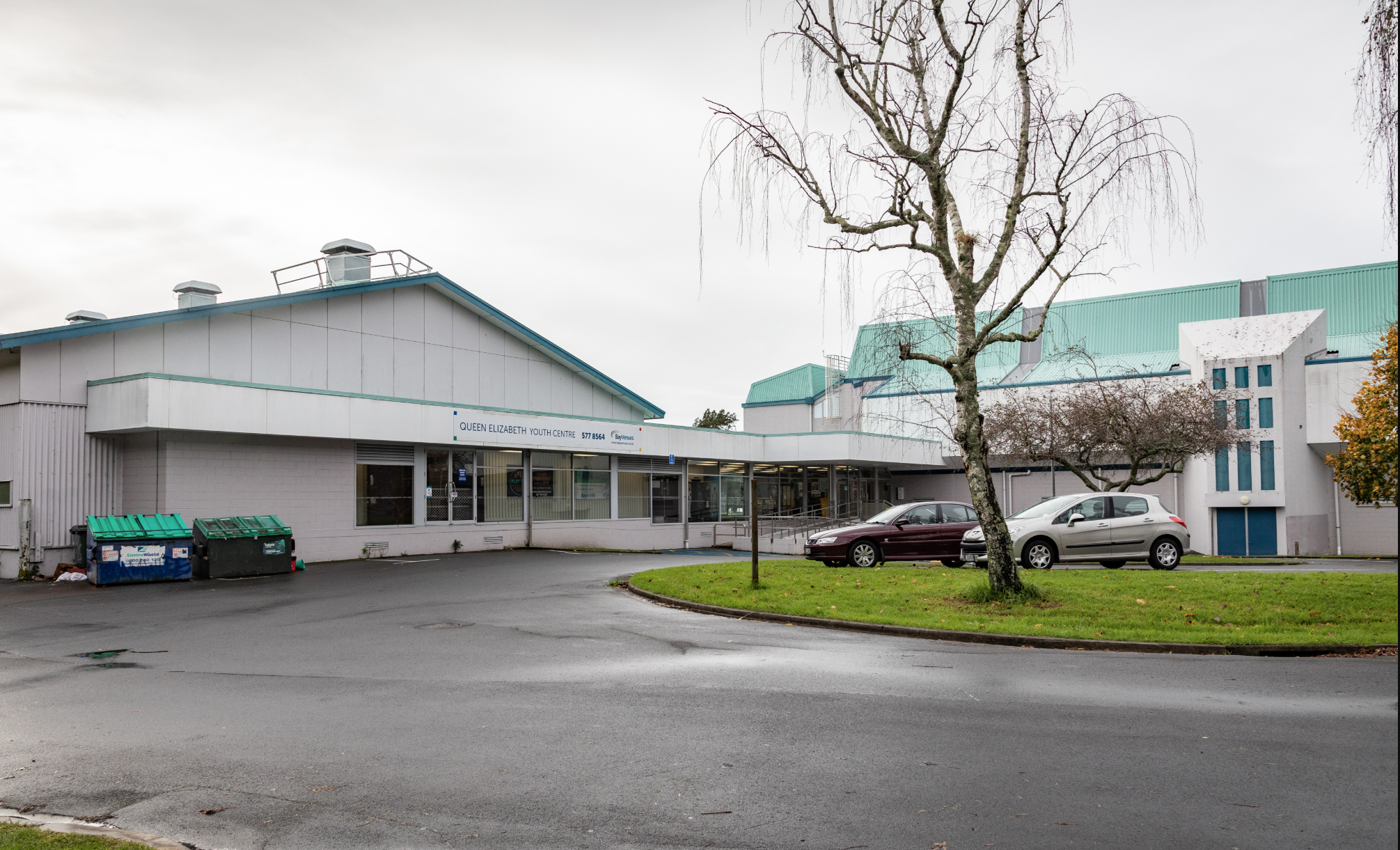 Level asphalt forecourt leads to the glass entrance of the Queen Elizabeth Youth Centre. A ramp with handrails is visible. Grey building with a green roof and clear signage on the fascia.