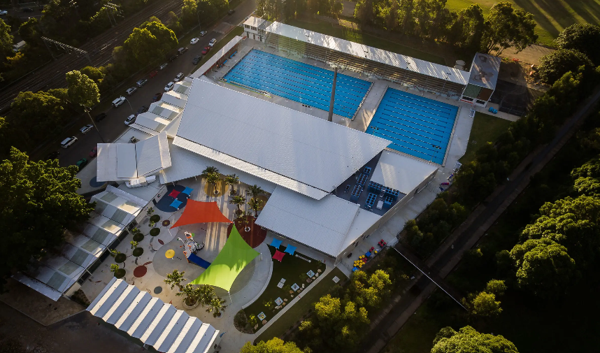 Level concrete pathways surround two outdoor pools and a children’s play area with orange and lime shade sails. Wide flat surfaces connect building entrances and street-level parking.